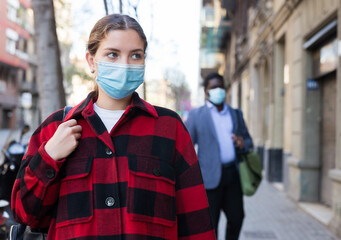 Young woman in disposable face mask walking along city street. Concept of Covid 19 virus spread...