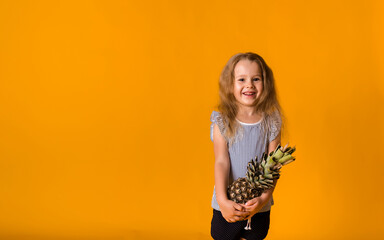 cheerful little blonde girl in a striped T-shirt stands and holds a pineapple on a yellow background with a copy of the space. Healthy fruits