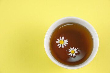 white cup with chamomile tea on a yellow background and with chamomille flowers floating on the infusion