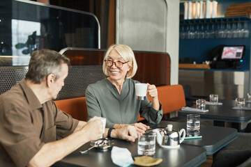 Positive senior Caucasian couple sitting at table with teapot and drinking tea while chatting in cafe