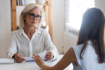 Pensive middle-aged businesswoman listen to young employee or job candidate at interview in office....