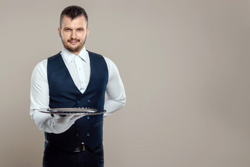 Handsome male waiter, white shirt, holds a silver tray, hand behind his back. Concept of wait staff serving customers in a restaurant.