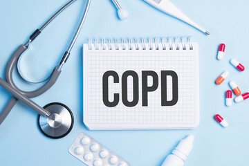 The word copd written on a white notepad on a blue background near a stethoscope, syringe, electronic thermometer and pills. Medical concept