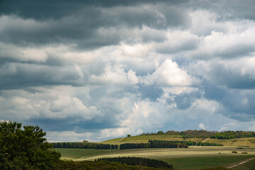 sunshine bursts through heavy blue and grey rain cloud on to a landscape with open grassland...