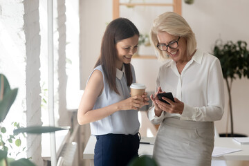Happy diverse female colleagues have fun drink coffee at break in office, use modern cellphone gadget together. Smiling coworkers watch video on smartphone, enjoy free time at workplace.