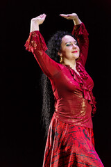 Gypsy woman with long black hair dances with arms with sleeves in a red dress on a black background.