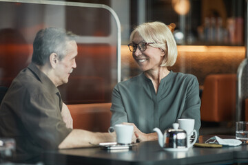 Happy senior Caucasian woman in eyeglasses sitting at table and holding hand of boyfriend while looking with love at him in cafe