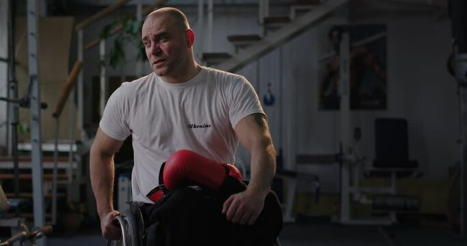 A Paralympian man with defective legs sits in a wheelchair in an empty gym sadly focused on the camera holding boxing gloves