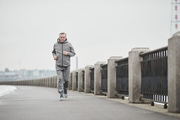 Purposeful senior Caucasian man in gray sport suit swinging arms while running alone in cold morning