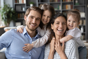 Portrait of smiling young parents have fun play with excited little preschooler daughters at home. Happy Caucasian mom and dad with small girls children hug enjoy leisure weekend. Family concept.