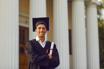 Young smiling university graduate in traditional costume standing and holding diploma in hand over...
