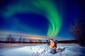 Concept photo tour to arctic travel, photographer man with camera and tripod photographs aurora borealis, northern lights green - obrazy, fototapety, plakaty
