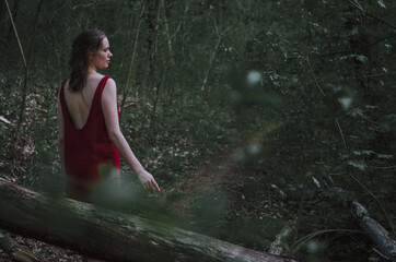 Fototapeta premium Atmospheric artistic portrait of woman in burgundy red backless linen dress walking in an enchanted forest 