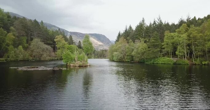4k aerial footage of Glencoe Lochan. Flying low over the water of the lochan past a small island with surrounding forest and mountains in background. Cloudy day in springtime.