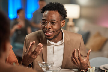 Portrait of elegant African-American man talking to friends at dinner table and gesturing actively