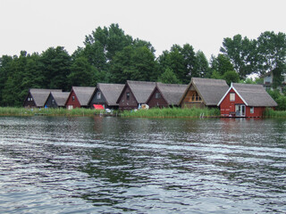 Fototapeta na wymiar Some colorful fishermen houses on the inland lake Müritz in Mecklenburg Vorpommern. Reeds grow in front of the wooden houses and some jetties reach into the lake. Behind the thatched roofs are trees