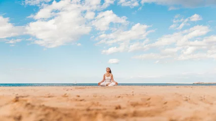 Gordijnen Senior woman in lotus pose sitting on the sand - Yoga at beach - Calm and meditation concept  © Davide Angelini
