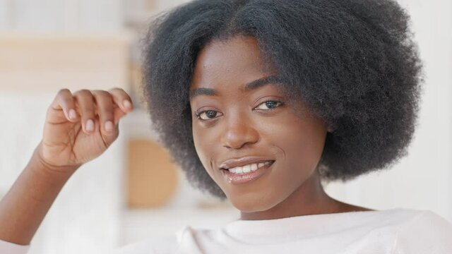 Portrait afro american ethnic beautiful curly woman african girl looking at camera twists curl on finger flirts posing smiling at home, close-up female face with perfect dark skin with natural make-up