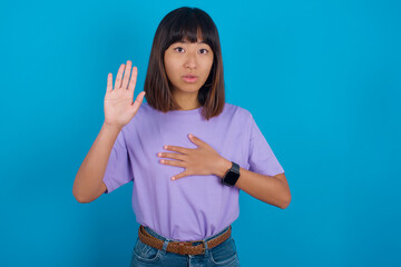 young beautiful asian woman wearing purple t-shirt against blue wall Swearing with hand on chest and open palm, making a loyalty promise oath