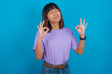 young beautiful asian woman wearing purple t-shirt against blue wall relax and smiling with eyes closed doing meditation gesture with fingers. Yoga concept.