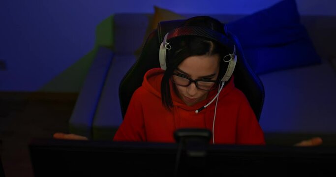 Beautiful girl in headphones and glasses sits in a dark room with backlight and broadcasts online, she speaks and gestures. Girl blogger is holding a conference on the network, modern technologies. 4k