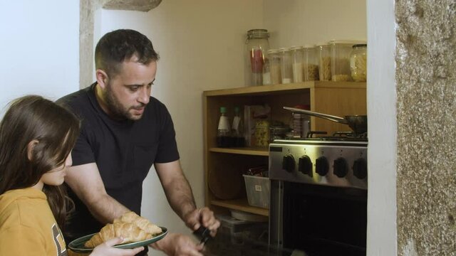 Solicitous father learning daughter how to use oven. Cheerful Caucasian dad and daughter putting plate of croissants in oven and lightning it together. Fatherhood, cooking concept.