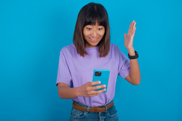 young beautiful asian woman wearing purple t-shirt against blue wall holding in hands cell reading browsing news