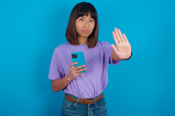 young beautiful asian woman wearing purple t-shirt against blue wall using and texting with smartphone with open hand doing stop sign with serious and confident expression, defense gesture