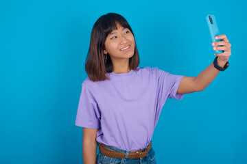 young beautiful asian woman wearing purple t-shirt against blue wall smiling and taking a selfie...