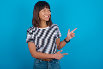 Optimistic young beautiful asian woman wearing stripped t-shirt against blue wall points with both hands and  looking at empty space.