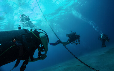 Close up photo of three divers swimming after each other close to anchor chain in blue water. Light...