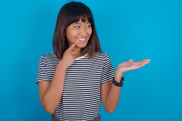 Fototapeta premium Positive young beautiful asian woman wearing stripped t-shirt against blue wall advert promo touch finger teeth