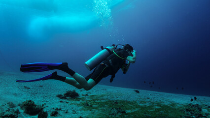 Young diver adjusts goggles just at beginning of diving session. Murky water, sandy bottom, boat silhouette at the water surface, anchor chain at left background