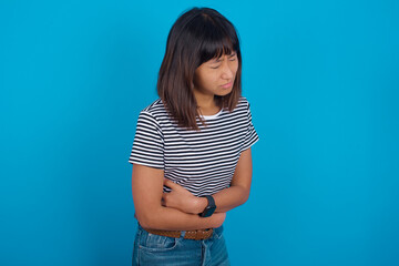 young beautiful asian woman wearing stripped t-shirt against blue wall got stomachache