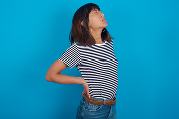 young beautiful asian woman wearing stripped t-shirt against blue wall got back pain