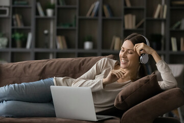 Convenient communication. Joyful young woman lie on couch before laptop in cordless earphones on head talk by video call. Teen female in headset use pc to study english online at easy language course