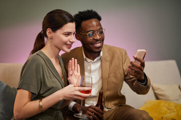 Portrait of smiling couple waving to smartphone screen while celebrating online via video chat in home setting
