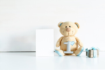 Little teddy bear toy with gift box, digit one on white table with copy space. Baby shower, accessories, stuff, present for boy child first year happy birthday, first newborn party background