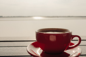 Red cup of hot latte coffee on wooden table at sunrise.