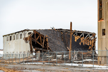 A dilapidated building made of metal and wood. The flat roof is tar and pitch with a metal fascia. The external wall of the building is yellow corrugated metal. There's a large opening with plywood. 