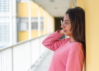 A pensive Latin woman standing on the hallway wearing glasses, with long dark hair, in a pink sweater