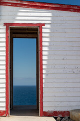 Fototapeta premium The entrance to a small white and red wooden building, which has two doors. Both doors are open allowing a view through the narrow woodshed. On the far side is the blue ocean with blue sky framed. 