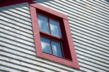 A bright red double hung vintage window with four glass panes. The residential window is on the exterior of a narrow horizontal white wooden clapboard siding wall. The clouds are reflecting in window.