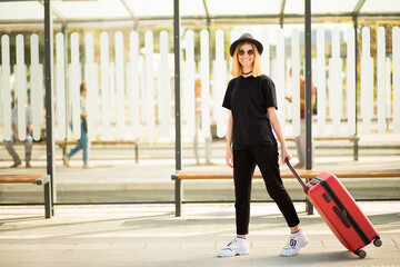 Young casual female traveler in total black and sunglasses walking and pulling a red wheeled luggage. Concept of travel. Copy space