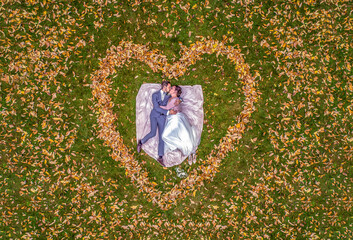 Aerial view of marriage couple lying in a shape of heart made of fall Autumn leafes woods drone...