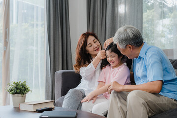 Asian grandparents kiss granddaughter cheek at home. Senior Chinese, old generation, grandfather and grandmother using family time relax with young girl kid lying on sofa in living room concept.