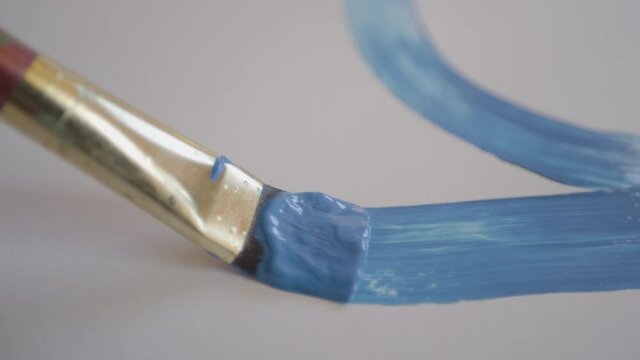 CLOSE UP, DOF: Detailed shot of a brush drawing a curved line with light blue paint as unrecognizable painter drags their fine brush across the blank canvas. Artist paints with blue acrylic paint