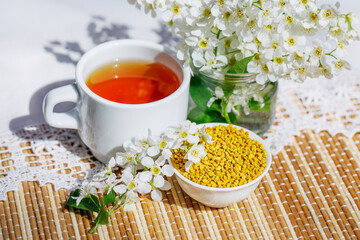 A bowl of pollen and aromatic herbal tea. Still life in a rustic style with a branch and flowers of a bird cherry and with a white knitted napkin. E