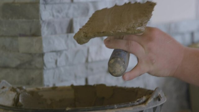 SLOW MOTION, CLOSE UP, DOF: Tiler uses a trowel to apply cement to a wall while laying tiles. Contractor applies mortar on the bare wall while laying natural stone tiles on the living room walls.