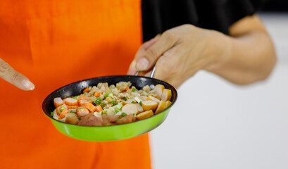 Tasty healthy food of hotdog, hams, crab stick on green flying pan shown by mom on orange apron that learned to cook for kid and family at home kitchen